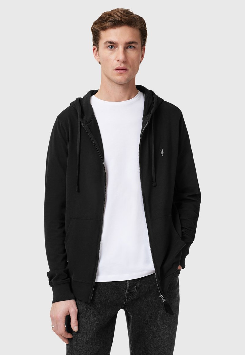 Felpa nera con zip e cappuccio regolabile, tasche frontali e un piccolo logo sul petto, indossata su una maglietta bianca e abbinata a jeans scuri.
