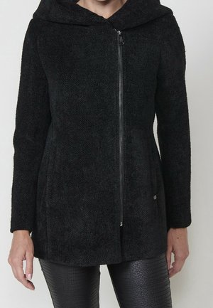 Femme portant une veste à capuche noire texturée avec fermeture éclair et un pantalon noir en similicuir à motif crocodile, debout devant un fond uni.