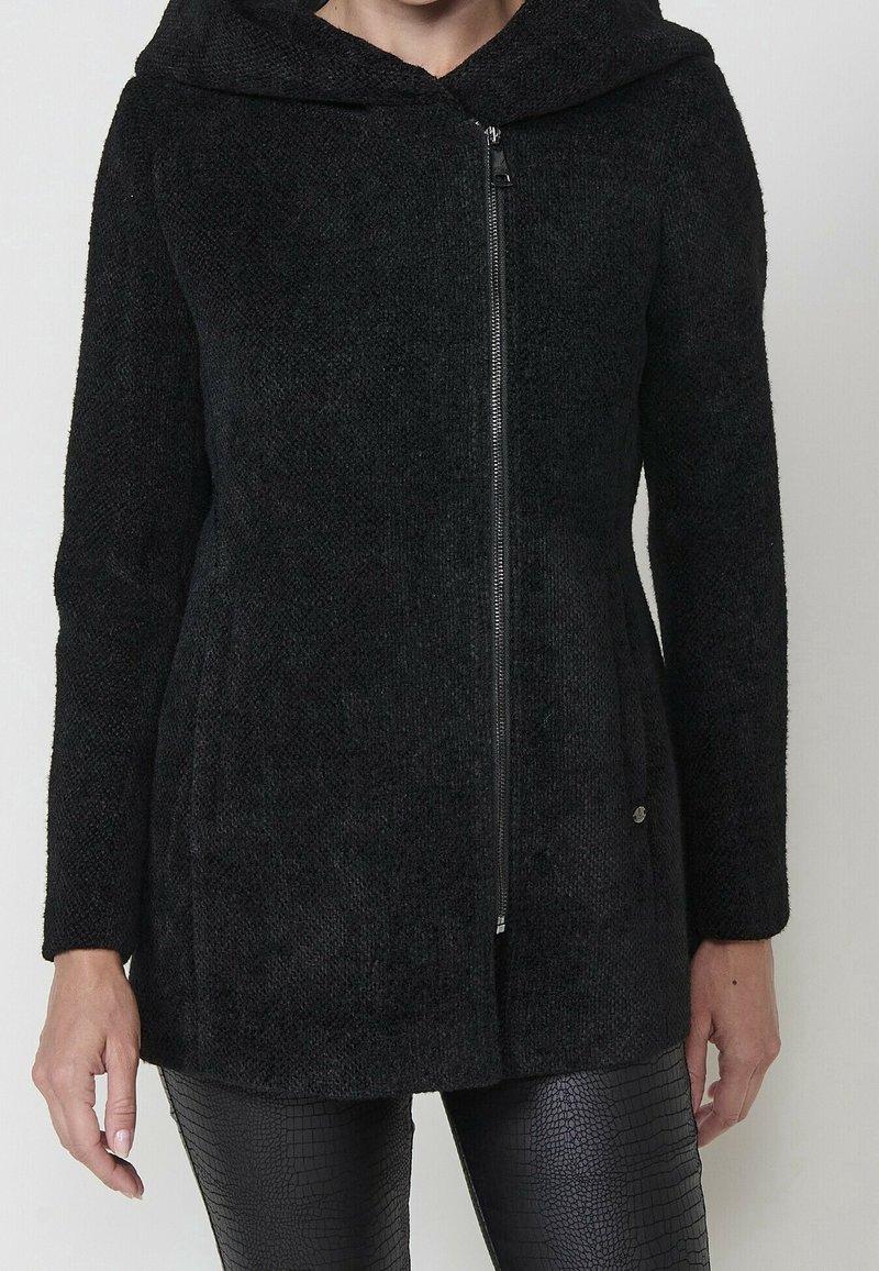 Femme portant une veste à capuche noire texturée avec fermeture éclair et un pantalon noir en similicuir à motif crocodile, debout devant un fond uni.