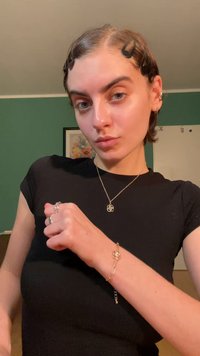 Auksinis apyrankė su gėlių motyvu, dėvima ant riešo. Modelis vilki prigludusią juodą marškinių, demonstruodama kelis žiedus ir minimalius papuošalus.