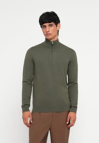 Olive green pullover sweater with high collar and quarter zip, ribbed cuffs and hem, soft texture, fitted design, worn with brown trousers.