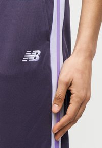 Κοντινό πλάνο ενός χεριού που ακουμπάει δίπλα σε σκούρο μοβ αθλητικό παντελόνι με λευκές και ανοιχτό μοβ ρίγες και λευκό λογότυπο New Balance.