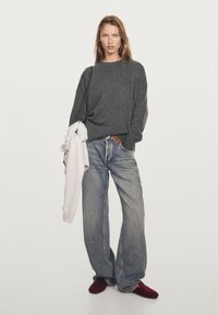 Frau mit einem dunkelgrauen Pullover, lockeren blauen Jeans, roten Schuhen, die einen weißen Pullover über ihre Schulter hält, vor einem weißen Hintergrund stehend.