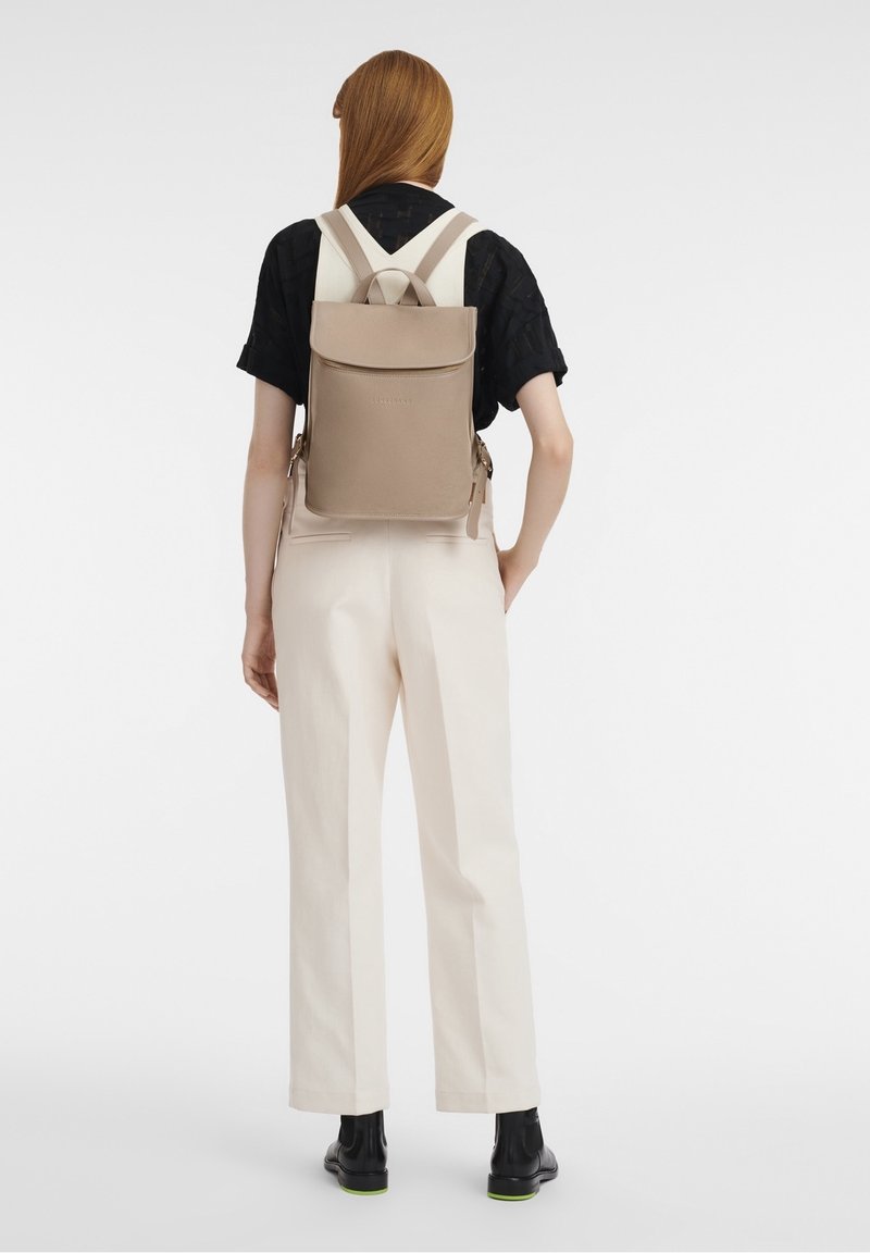 Femme portant un chemisier noir et un pantalon blanc, debout avec un sac à dos en cuir beige sur fond blanc.