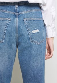 Osoba v světle modrých džínách s malou dírou na pravé zadní kapse a bílou košili s dlouhým rukávem viditelnou na pravé paži.