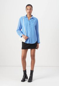 Camicia azzurra chiara a maniche lunghe, abbinata a una mini gonna nera e stivaletti neri alla caviglia. Il modello tiene in mano una piccola borsa a tracolla bianca.
