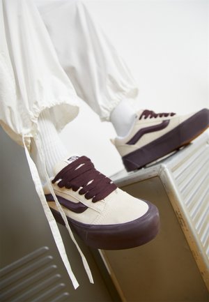 Personne portant des baskets crème et marron foncé avec des semelles épaisses et des chaussettes blanches, assise avec les pieds reposant sur une surface métallique.