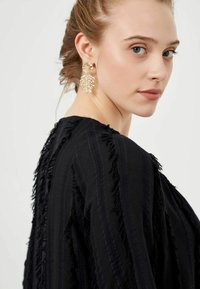 Zwarte kleding met rafelige randen en getextureerde strepen, gedragen met sprankelende gouden bladvormige oorbellen. Model toont een zijaanzicht.
