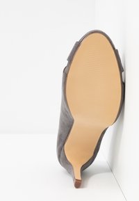 Šedá semišová lodička na vysokém podpatku s špičatou špičkou, s hladkou béžovou podrážkou a texturovaným gumovým detailem blízko podpatku.
