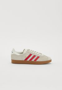 Adidași din suede bej cu dungi roșii și albastre; talpă din cauciuc; vârf rotund; accente albe și etichetă de text aurie pe lateral.