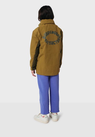 Person in einer olivgrünen Jacke mit kreisförmigem Text auf dem Rücken, lila Hosen und weißen Sneakers, vor einem schlichten hellen Hintergrund stehend.