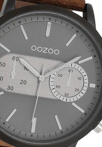 Montre analogique ronde avec un cadran gris, des index argentés et deux sous-cadrans. Elle possède un boîtier noir et un bracelet en cuir brun.