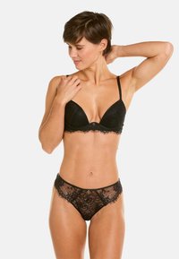 Soutien-gorge en dentelle noire avec un décolleté plongeant et des bords festonnés, associé à une culotte en dentelle transparente assortie ornée de motifs floraux et de matériel minimal.