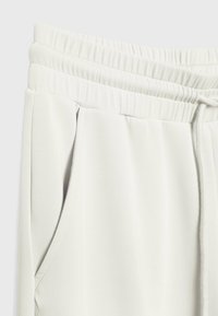 Zbliżenie białych spodni dresowych pokazujące elastyczny pas, sznurek do zawiązywania oraz szczegóły bocznej kieszeni na jednolitym tle.