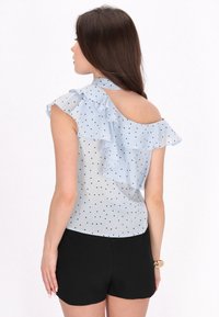 Bluză de culoare albastru deschis cu un model punctat, cu volane stratificate pe umeri și spate deschis. Asortată cu pantaloni scurți negri.