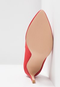 Chaussure à talon haut rouge avec un bout pointu, un dessus en textile lisse, et un talon fin. La semelle est d'un beige clair avec une texture nervurée.