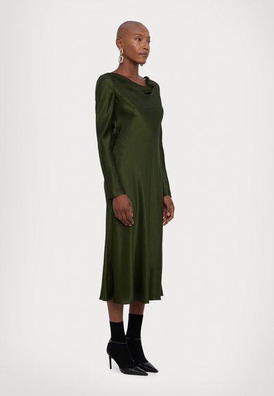 Satin groene jurk met lange mouwen, een kraaghals en zijsplitten. Gecombineerd met zwarte enkellaarsjes en aansluitende sokken.