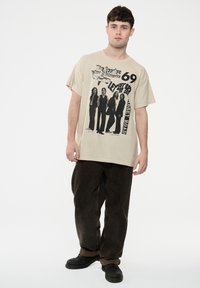 Beige graphic t-shirt featuring a Beatles print with text, short sleeves, relaxed fit, paired with brown pants and black shoes.
