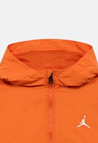 Πορτοκαλί μπουφάν με κουκούλα επενδεδυμένη με διχτυωτό υλικό, μισό φερμουάρ στο μπροστινό μέρος και λευκό λογότυπο Jumpman. Το υλικό φαίνεται ελαφρύ και λείο.