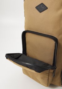 Tan fabric backpack with a textured exterior, featuring a front black zippered pocket and a logo patch at the top.