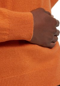 Maglione arancione lavorato a maglia con polsini e orlo a coste. Texture morbida e design senza cuciture, con un primo piano di una mano appoggiata sul tessuto.