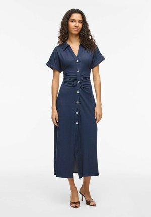 Kvinde med krøllet hår iført en navy knap-foran midi-kjole med korte ærmer og rynket talje, stående i brune åbne hæle.