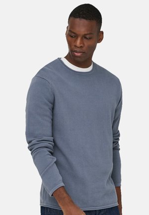 Mies pukeutuneena vaaleansiniseen pitkähihnaiseen, tekstuuriin neulottuun neulepaitaan, valkoisen aluspaidan päällä, katse käännettynä alas vasemmalle yksinkertaisen valkoisen taustan edessä.