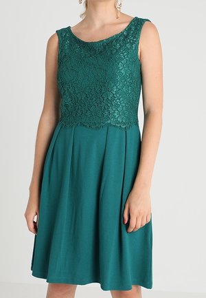 Robe sans manches bleu sarcelle avec corsage en dentelle florale et jupe unie portée par une personne debout devant un fond uni.