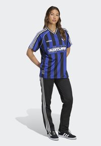 Žena stojící se založenýma rukama za zády, oblečená v modro-černém pruhovaném sportovním dresu Adidas, černých sportovních kalhotách Adidas a černých teniskách.