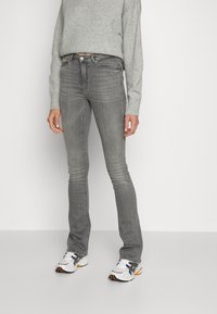 Jeans cinzas justos com uma ligeira boca de sino, apresentando uma cintura média e uma textura suavemente desbotada. Combinados com um suéter cinza claro e ténis desportivos.