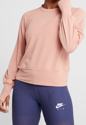 Persoon draagt een lichtroze longsleeve top en donkerblauwe Nike Air legging, met de linkerarm verhoogd bij het gezicht.