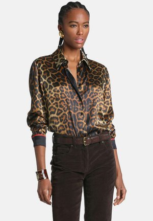 Femme aux cheveux tressés portant une chemise en satin à imprimé léopard, un pantalon marron foncé avec ceinture, de grandes boucles d'oreilles dorées et un bracelet marron.