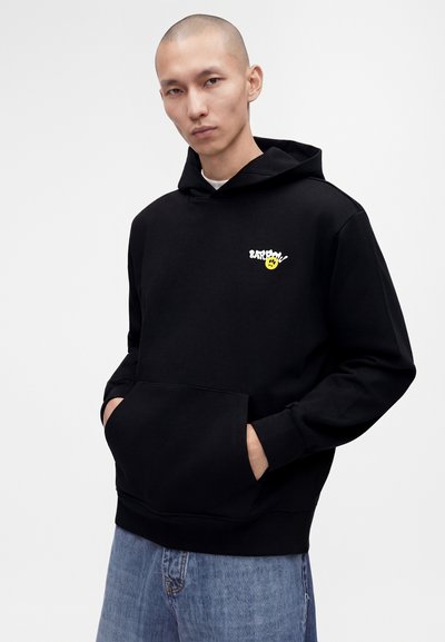 Homme portant un sweat à capuche noir avec un petit motif blanc et jaune sur la poitrine et un jean bleu, mains dans la poche avant, fond neutre.