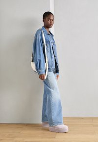 Veste en denim bleue, coupe décontractée avec grandes poches. Jeans évasés bleu clair avec fentes latérales. Chaussures platforms rose clair et sac à main blanc.