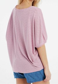 Nainen, jolla on yllään löysä vaaleanpunainen chevron-kuviollinen paita kyynärtaipeeseen ulottuvilla hihoilla ja siniset farkkushortsit, nähtynä takaapäin.