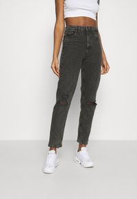 Kvinna i högmidjade svarta slitna jeans, vita sneakers och en ärmlös vit crop top som står mot en neutral bakgrund.