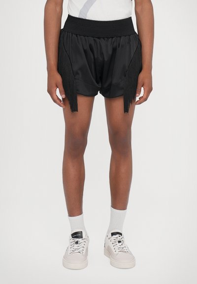Schwarze Satinhorts mit fransigen Akzenten an den Seiten, elastischem Bund und lässigem Schnitt. Kombiniert mit hellen Sneakers und Socken.