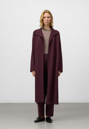 Frau steht gegen eine weiße Wand und trägt einen langen, pflaumenfarbenen Mantel, passende Hosen, einen taupefarbenen Pullover, schwarze Slipper und goldene Halsketten.