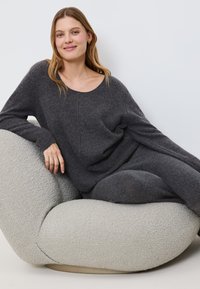 Frau mit langen blonden Haaren, die einen dunkelgrauen Strickpullover trägt und auf einem strukturierten hellgrauen modernen Lounge-Stuhl vor einem weißen Hintergrund entspannt.
