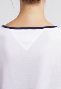 Valkoinen T-paita, jossa on tummansininen kaula-aukko ja kolmionmuotoinen yksityiskohta takana. Pehmeä materiaali ja rento malli.