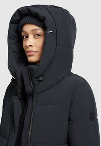 Chaqueta de plumas negra con una gran capucha, con textura acolchada, cierre de cremallera y detalles de hardware grabados.