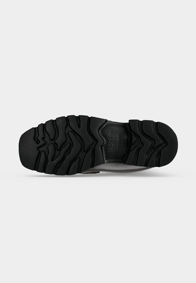 Semelle d'une chaussure en caoutchouc noire avec des motifs de sculpture profonds et un patch rectangulaire avec un texte embossé au centre.