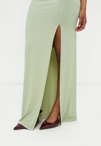 Jupe maxi vert clair avec une fente haute à l'avant, portée avec des escarpins slingback à bout pointu rouge foncé sur les jambes d'un mannequin.