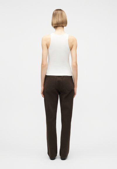 Άτομο με κοντά ξανθά μαλλιά που φοράει λευκό αμάνικο τοπ και σκούρα καφέ παντελόνια ίσιας γραμμής, στέκεται γυρισμένο από την πλάτη σε απλό φόντο.