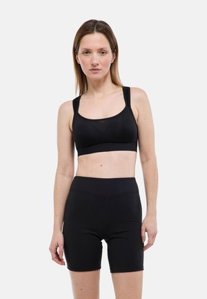 Femme portant un soutien-gorge de sport noir et un short cycliste noir taille haute, debout devant un fond blanc uni.