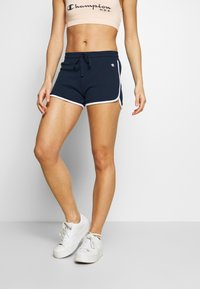Mörkblå sportshorts med vita sidsömmar och dragsko i midjan, i kombination med en beige Champion crop top och vita sneakers.