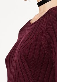 Burgundy neulepaita, jossa on pystysuora ribbikuvio, leveä pääntie ja sileä viimeistely. Korostettu mustalla kaulaketjulla.