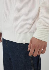 Pull en tricot blanc et rose clair avec des rayures verticales texturées. Présente un ourlet côtelé et des poignets, ainsi qu'une petite étiquette en bas.