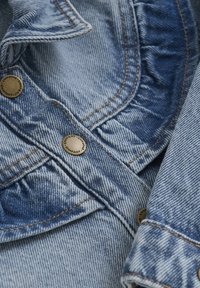 Ljusblå denimjacka med volangdetalj, mässingsknappar och kontrasterande orange sömmar. Mjuk textur, klassisk design.