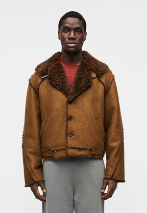 Καφέ δερμάτινο μπουφάν με μεγάλος γούνινος γιακάς, τρία μαύρα κουμπιά και πλαϊνές τσέπες. Σχεδιασμός με υφή, κοντός με ξέφτια στα άκρα.
